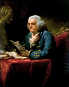An old man in Revolutionary-era wig, blue with gold jacket, and white ruffled shirt sits in a red satin chair at a small table in red satin. He holds a slightly crumpled manuscript in one hand, which he reads, glasses over noses, as he rests his soft chin on the thumb of his other hand.