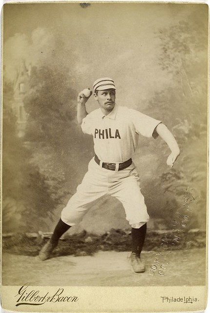 An early-1900s baseball player from team Philly winds back to throw. Stern eyes and a broad and full but not-quite handlebar mustache.